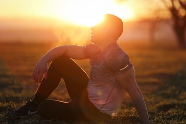 Muscular man sits in rural field at sunset, exhaling visible breath.